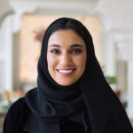 Indoor portrait of woman in traditional black abaya and hijab smiling at camera while standing in hotel lobby with Islamic architectural design.